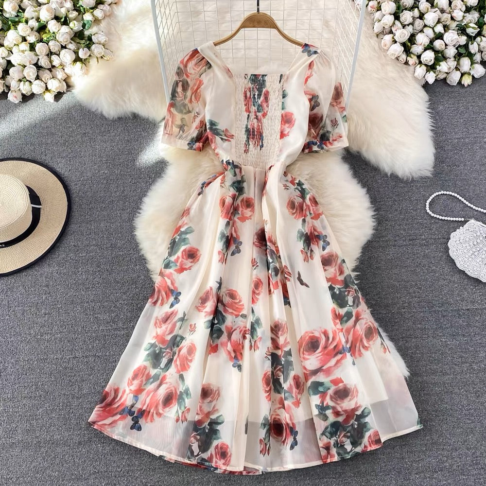 A floral print maxi dress displayed on a white furry surface with a pleated and ruched design, beige in color, with accessories like a hat and a handbag in the background.